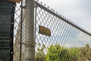 Chain-link fence with a metal post and a small yellow sign, with trees in the background.