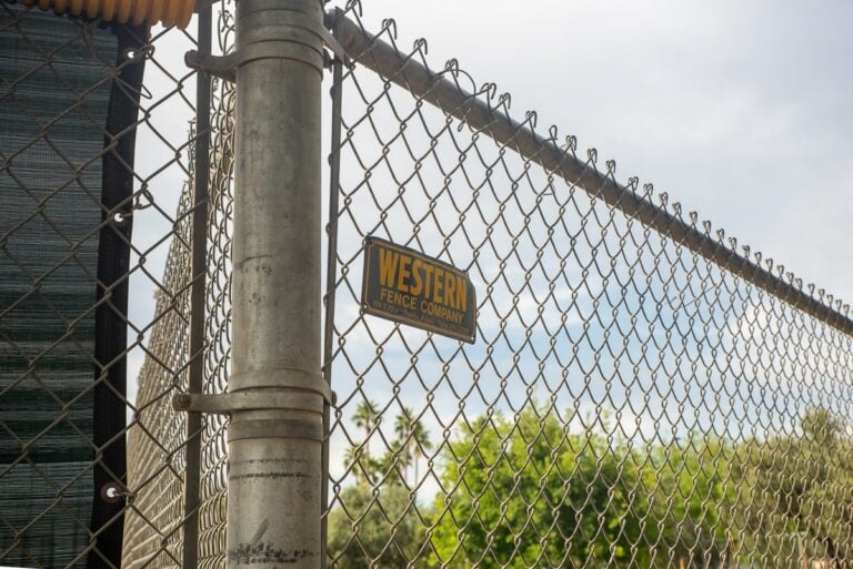 Chain-link fence with a metal post and a small yellow sign, with trees in the background.