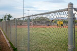 Chain-link fence runs along a baseball field with a yellow Western sign in the outfield under a blue sky.