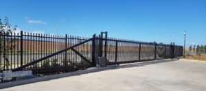 Black metal gate and fence across a paved driveway in a rural landscape under a clear blue sky.