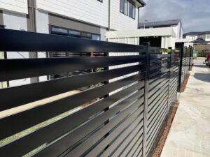 A black metal fence with horizontal slats runs along a sidewalk beside modern residential buildings.