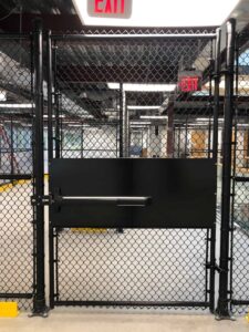 Indoor gym cage area with a black solid-panel gate set in chain-link fencing and an overhead exit sign.