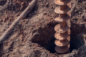 A metal auger drill bit is embedded in sandy soil, creating a hole.