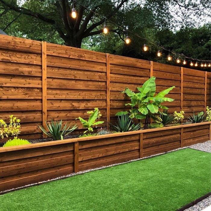 Backyard with a tall wooden fence, a raised planter bed with plants, string lights overhead, and a green artificial lawn.