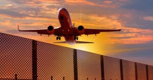 A red airplane with landing gear down approaches for landing over a chain-link fence at sunset.
