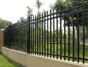 Black wrought-iron fence with vertical bars and pointed tops sits on a low brick wall beside a grassy park with trees.