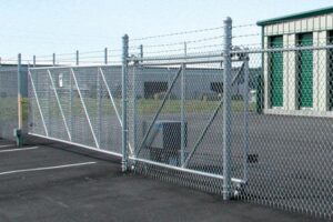 Chain-link fence gate across a paved lot with a small green building in the background.