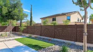 Backyard with a tall brown privacy fence, gravel landscaping, a small green lawn, and a beige two-story house visible in the background under a bright blue sky.