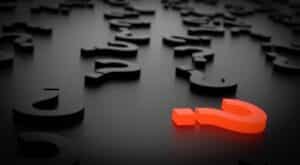 A bright orange question mark lies among scattered black punctuation marks on a dark, reflective surface.