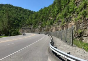 A winding two-lane road curves along a rocky hillside with a metal guardrail and chain-link fence, green trees lining the slope, and a car visible in the distance.
