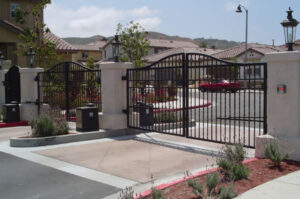 Gated residential entrance with curved black iron gates between stone pillars, a guard booth on the left, and landscaped shrubs with houses visible in the background.