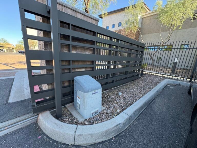 Dark horizontal-slat fence beside a gravel bed with a gray utility box and a curved concrete curb in a parking area.