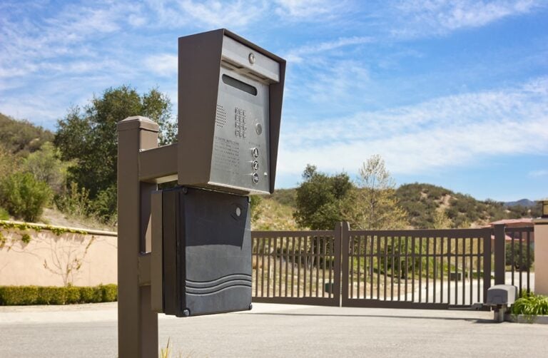 Gray outdoor mailbox mounted on a wooden post beside a metal gate with desert hills and a blue sky in the background.