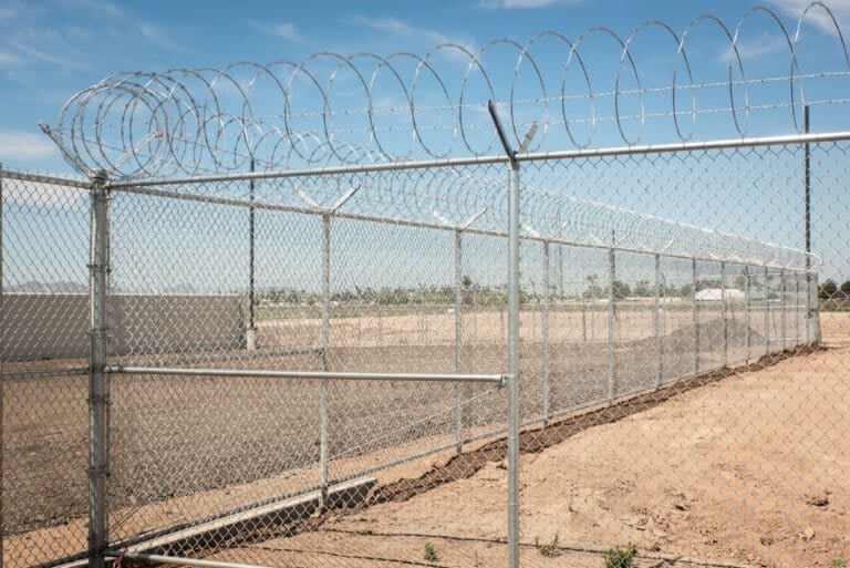 Chain-link security fence topped with razor wire extends across a dry, desert-like landscape.
