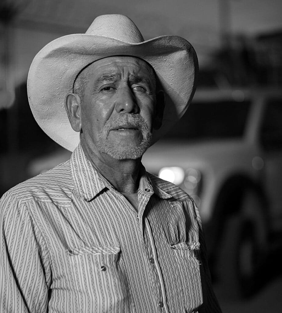 An elderly man wearing a wide-brimmed cowboy hat and striped shirt stands outdoors with a blurred vehicle in the background.