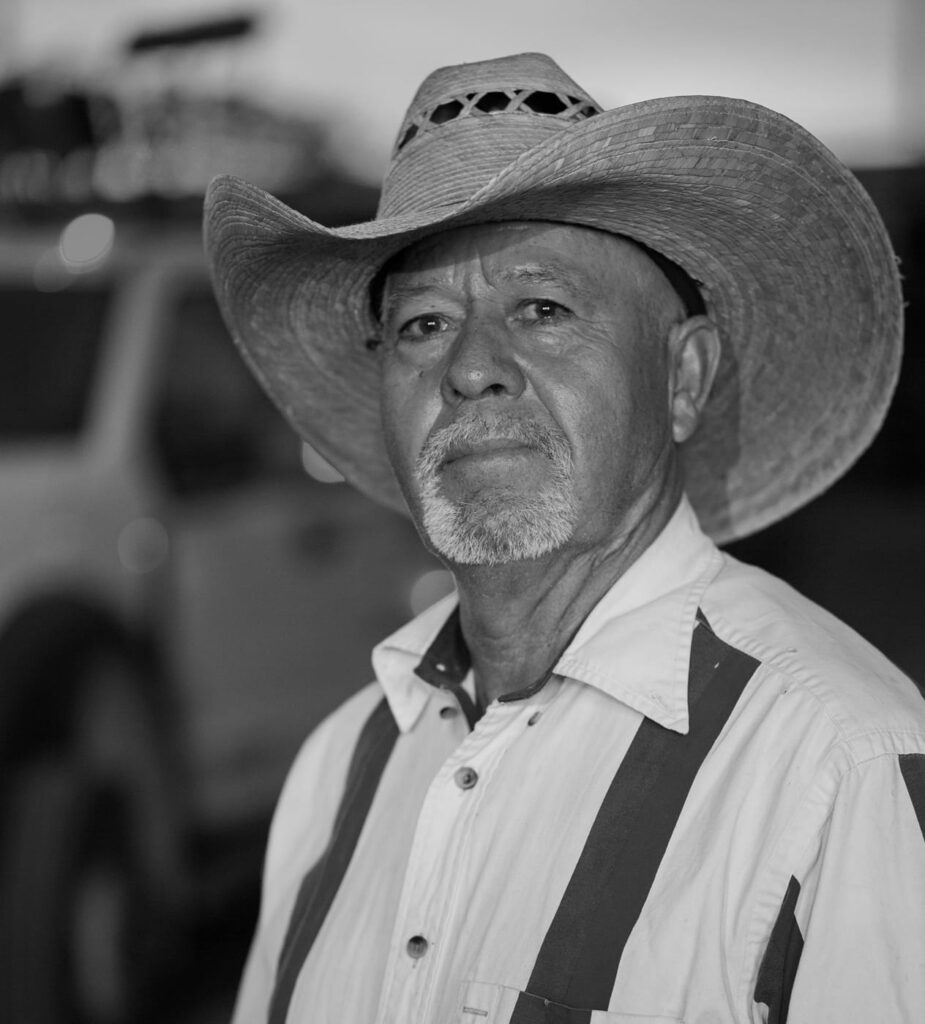 Black-and-white portrait of an older man wearing a cowboy hat, a white shirt, and suspenders, looking at the camera.