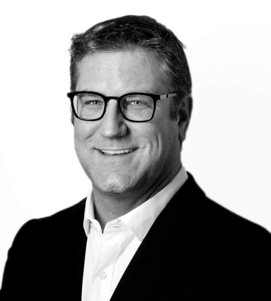 Smiling man wearing glasses, a white shirt, and a dark blazer against a white background.