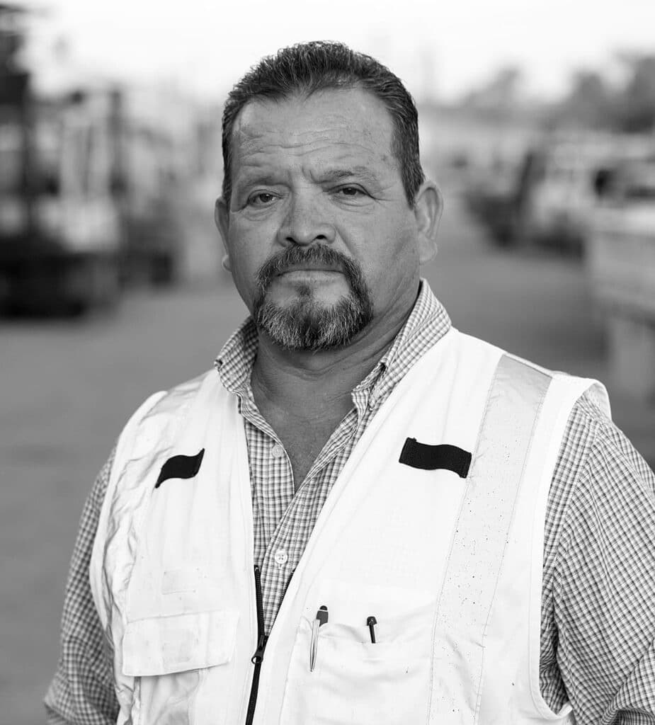 A middle-aged man wearing a white safety vest stands outdoors on a street.