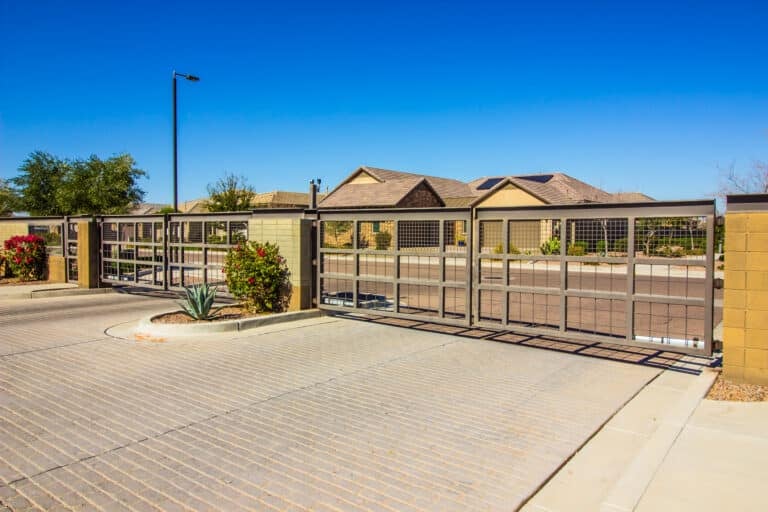 Gated residential community entrance with brick pillars, a metal gate, and a paved driveway leading to houses under a clear blue sky.