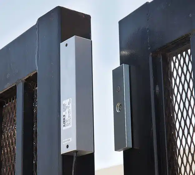 Close-up of an electric strike lock mounted on a metal gate with a mesh panel.