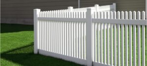 White vinyl picket fence along a green lawn with a house in the background.
